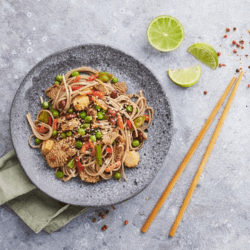 A dish of pasta infused with beef cutlets, sausage slices, peas, and other vegetables served on a grey dish with lime slices and chopsticks displayed on the side