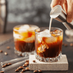 Two glasses of Iced Kahlua coffee with ice and cream showing a hand pouring more cream in the first glass